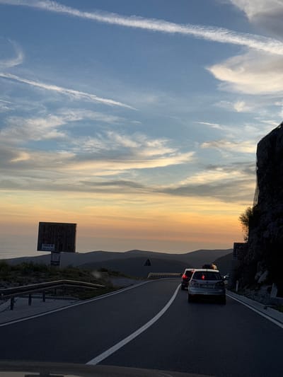 The image depicts a sunset scene on a mountain road in Stanišići, Montenegro. The sky is filled with a gradient of colors, transitioning from a pale blue at the top to hues of orange and yellow near the horizon, with scattered white clouds. A dark, rugged cliff face dominates the right side of the frame. The road, paved with asphalt, curves to the left and is marked with a white line. Several cars are visible on the road, appearing to be in traffic. On the left side of the road, a metal guardrail separates the road from a grassy slope. Further back on the left, a large, weathered billboard stands against the backdrop of rolling hills and the distant sea. The overall impression is one of a serene, yet slightly congested, mountain drive during twilight.