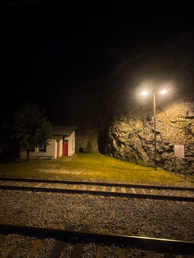 This image depicts a nighttime scene at a train station in Lopate, Montenegro. In the foreground, two sets of train tracks are visible, laid on a bed of dark gravel. The tracks recede into the darkness. Behind the tracks, a patch of illuminated grass slopes upwards towards a small, white building with a red door and a metal roof. A tree stands to the left of the building. Further back, a steep, rocky cliff face dominates the background, illuminated by a tall street lamp with two light fixtures. The lamp casts a bright glow on the rock and the grassy slope, creating stark shadows. The overall ambiance is one of isolation and quiet. There are no people visible in the image.