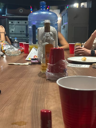 A table displays an array of objects: a large water bottle, a clear liquor bottle, stacks of red plastic cups, a plate with food, an e-cigarette, and assorted food wrappers. People's arms and hands are visible at different points around the table. The setting appears to be an outdoor gathering, judging from the blurred background featuring what might be a building.