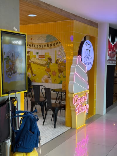 The image depicts the exterior of a Roti Boy and Cool Boy establishment in Pajang, Indonesia. The storefront features bright yellow tiled walls and a mural with cartoon figures. A large neon sign in the shape of an ice cream cone with the words "Cool Boy" is prominently displayed, casting a pink glow on the white tiled floor. To the left, a digital display board with a yellow frame shows images of food items. In the foreground, a blue backpack is slung over a luggage stand, and a yellow suitcase is visible beneath it. Several black metal chairs and tables are set up outside the shop. The overall ambiance is colorful and inviting, suggesting a popular food and beverage spot.