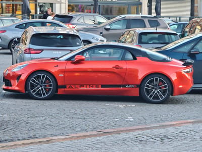 A vibrant red Alpine A110S sports car is the focal point of this image, parked on a cobblestone street in Ypres, Belgium. The car features a sleek design, a contrasting black roof, and stylish multi-spoke wheels with red brake calipers. Black lettering along its side proudly displays "ALPINE" and "A110S."
Behind the Alpine, a collection of other vehicles is parked, including a silver Kia Ceed hatchback. On the rear window of the Kia, text is visible, identifying the car as a "KIA CEED" and featuring a sticker with "Auwerx Flevo" and "Kortrijk," reinforcing the Belgian location. Other cars, including various SUVs and sedans, are also visible in the densely packed parking area.
In the background, two individuals can be seen near a building with large glass windows. One person wears a dark hoodie, while the other is in a lighter-colored top. Partial text on the building's facade, possibly "roos parts h" and "Ayants P," suggests a commercial establishment. The cobblestone pavement indicates an urban setting, typical of historic European towns. The lighting is diffused, suggesting a daytime scene, possibly overcast or during the late afternoon. The overall atmosphere is that of a regular day in a busy city center.