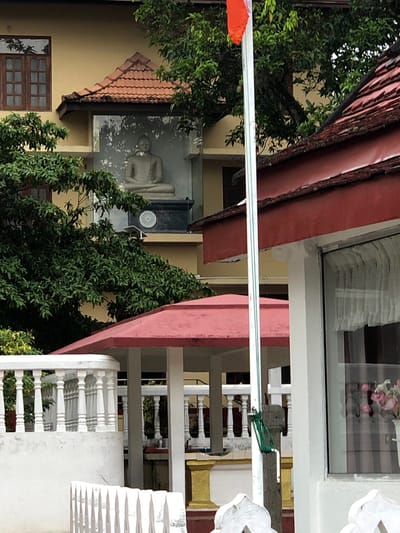An outdoor shot shows a multi-story building with a Buddha statue inside a glass-enclosed alcove on an upper floor. The building's exterior is painted yellow, with a red-tiled roof. To the right, another building with a similar red roof is partially visible, featuring a window with sheer curtains. A white railing with balusters runs along the foreground. Green foliage from trees is abundant throughout the scene. The image appears to be taken during the daytime. The specific location in Sri Jayewardenepura Kotte, Sri Lanka, is not discernible from the visual information provided.