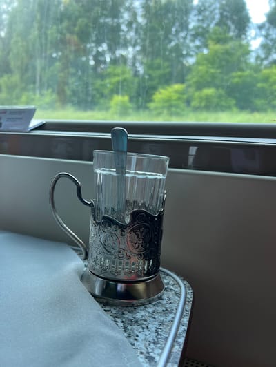 The image shows a glass of water in a metal cup holder with a spoon inside, placed on a granite-patterned surface next to a white tablecloth. In the background, through a window, a blurry green landscape of trees and foliage is visible, suggesting a moving vehicle, possibly a train, as indicated by the presence of the cup holder, a common item on Russian trains. The scene is likely set inside a train carriage traveling through the countryside near the city of Keramik, Russia, on a day with overcast skies. There are no people visible in the image.
