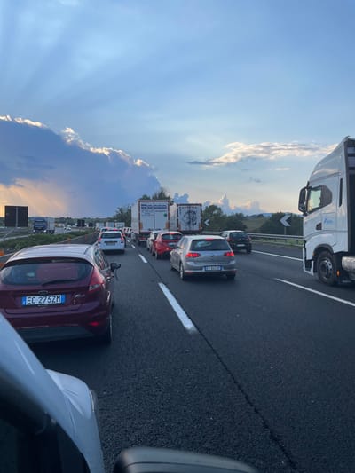 The image depicts a multi-lane highway experiencing heavy traffic congestion in the vicinity of Paliano, Italy. The scene is illuminated by late afternoon or early evening light, with crepuscular rays visible through a partly cloudy blue sky. Vehicles are stationary or moving slowly in two visible lanes.

In the immediate foreground, the lower portion of a white vehicle, from which the photograph was taken, is visible on the left, including part of a rearview mirror. Directly ahead in the left lane is a maroon Ford Fiesta hatchback bearing the Italian license plate EC 275ZM. In the right lane, a silver Volkswagen Golf with Italian license plate ET 845IP is positioned ahead of a red passenger car. Further down the highway, a white Iveco semi-truck with a "Falco" logo is partially visible on the far right. Multiple other commercial vehicles are present in the mid-ground, including two white trailer trucks, one identified with "ROCCO Trasporti" text and another displaying a dark animal head graphic.

The road surface is black asphalt, delineated by solid and broken white lane markings. The environment features green, undulating hills and scattered trees in the background to the right. A dark, square structure, likely a road sign gantry support, is visible on the distant left horizon. No individuals are visible outside of the vehicles.