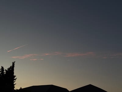 This media file captures a serene twilight scene in Mastaga, Azerbaijan. The primary subjects are the silhouettes of rooftops and trees against a dusky sky. The sky itself is a gradient of deep blue at the top, transitioning to a lighter, yellowish hue near the horizon, suggesting the last moments of daylight.
A notable detail is a thin, reddish contrail streaking diagonally across the upper left portion of the sky, indicating an aircraft's passage. Below this, a band of thin, pinkish clouds stretches horizontally, catching the fading sunlight. The silhouettes of several pointed rooftops, likely from houses or other buildings, form a dark band across the bottom of the frame. To the left of the rooftops, the dark, feathery silhouette of a tall, slender tree or a cluster of trees stands prominently, its shape detailed against the lighter sky.
The overall mood is peaceful and quiet, characteristic of dusk. The lighting suggests a time shortly after sunset. There are no visible people or text in the image. The location is identified as Mastaga, Azerbaijan, which provides a geographical context to the otherwise universal scene of a fading day.