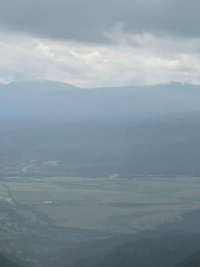 This is a high-angle or aerial photograph depicting a vast landscape, likely taken from an elevated position such as a mountain or an aircraft, in the region of Hristo Danovo, Bulgaria.
When viewed correctly oriented, the left and upper sections of the image are dominated by a partly cloudy sky, featuring prominent white and light gray cloud formations that stretch across the frame. Below the clouds, in the middle distance, a long, hazy mountain range extends horizontally, its slopes transitioning from lighter tones near the sky to darker, possibly forested, areas.
The lower and right portions of the image reveal expansive green plains, indicative of agricultural fields or pastures, interspersed with darker patches of vegetation or scattered trees. A small, distant white building or structure is faintly discernible within one of these fields in the lower right area.
The scene conveys a sense of peaceful expanse during daytime, with diffused natural light suggesting either an overcast sky or significant atmospheric haze. There are no people visible, no specific events taking place, and no discernible text. The image provides a panoramic view of Bulgaria's diverse topography, encompassing both mountains and agricultural lowlands.