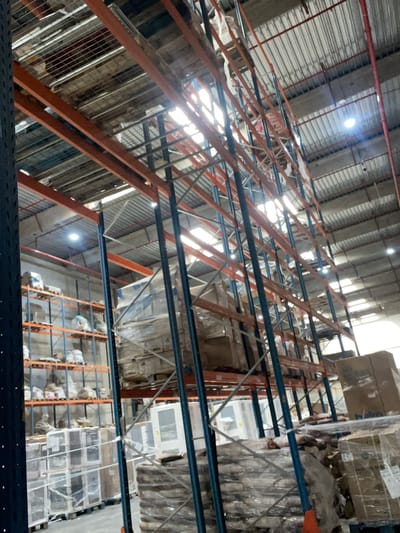 This is a long-shot, low-angle, eye-level, interior shot of a warehouse in Skhirat, Morocco. The warehouse has a high ceiling with corrugated metal roofing and visible lighting fixtures. Massive industrial metal shelving units, painted orange and blue, dominate the frame, extending vertically and horizontally to store a variety of goods. The shelves are laden with boxes, some wrapped in plastic, and other items stacked on pallets. The overall impression is one of a large-scale storage facility.
