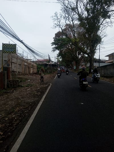 The image depicts a street scene in Sat Thay (East), Myanmar, under an overcast sky. A dark asphalt road, marked with a white solid line on its left edge, recedes into the distance. Multiple motorcycles are observed traveling away from the viewer on this road; at least four riders wearing helmets are visible. In the foreground on the left, a person, facing away from the camera, stands or walks on a dirt and debris-strewn shoulder, carrying a large bundle of branches or firewood on their back. Adjacent to this individual on the left is a construction site featuring concrete structural frames and wooden scaffolding. A yellow and black checkered sign reading "CLEAN GLOW CARWASH" is mounted on a pole above the construction. A dense cluster of overhead utility wires spans across the upper left quadrant. The right side of the road is lined with several large, mature trees with dense foliage and some visible epiphytes. Residential buildings are discernible in the background on both sides of the street.
