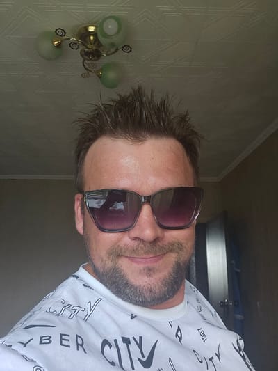 A selfie of a man wearing sunglasses and a white t-shirt with a repeating "CITY" pattern is taken from the chest up. The man has spiky brown hair and a beard. In the background, a ceiling light fixture with three green glass shades is visible, attached to a textured ceiling. The overall lighting suggests an indoor setting. The location of Таёжное, Russia is not evident from the image.