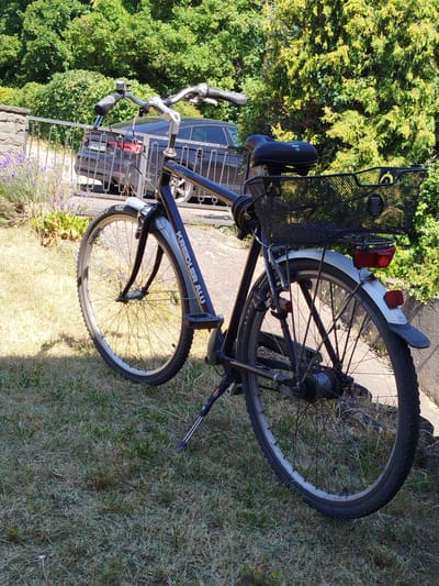 A dark-colored bicycle with a front basket and a rear rack is parked on a grassy area next to a paved path. The bicycle has a metal frame and appears to be a standard commuter or touring model. In the background, a dark sedan is visible behind a metal fence, and lush green trees and bushes provide a backdrop. A small stone wall or structure is also present to the left of the frame. The scene appears to be outdoors, likely in a residential or park setting. The license plate on the car reads "KT-ZS 114".