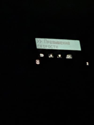The image displays a digital screen with a light green illuminated background against an otherwise dark setting. Prominently featured on the screen is Cyrillic text: ">> Превышение СКОРОСТИ", which translates to ">> Exceeding SPEED", indicating a speed violation. Below this text, four control icons are visible: a curved left arrow, an upward-pointing triangle, a downward-pointing triangle, and an "OK" button. Faint, illuminated numerals "1" and "2" are discernible in the lower left and lower right quadrants of the screen, respectively. The scene lacks visible people, external environment, or specific objects beyond the digital display, which is likely an in-vehicle interface or similar electronic device operating within the city of Voskresensk, Russia.