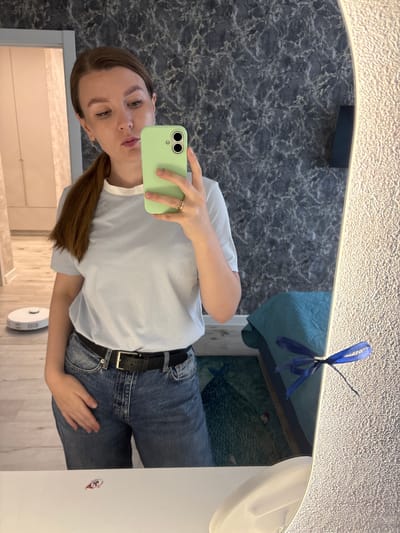 A young woman with brown hair tied in a ponytail is taking a selfie in a mirror. She is wearing a light blue t-shirt, blue jeans, and a black belt with a gold buckle. She is holding a mint green smartphone in her right hand. The background of the image shows a wall with a marble-like pattern, a doorway, and a robot vacuum cleaner on the floor. In the reflection of the mirror, a blue blanket and a piece of furniture are visible. The scene appears to be an indoor setting, likely a home, in the city of Izhevsk, Russia.