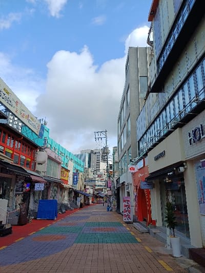 The image depicts a narrow pedestrian commercial street in 마포구 (Mapo-gu), South Korea, viewed from a low-angle perspective looking down its length. The sky above is partially cloudy blue.
The street is paved with multi-colored interlocking pavers in shades of blue, green, orange, and purple, forming a patterned path in the center and right. The ground on the left side is covered with a solid red surface.
Buildings on the left are predominantly older, multi-story commercial structures featuring red-framed windows and diverse storefronts with Korean signage. A blue tarp and a large brown woven bag are visible on the sidewalk in front of one shop. A large, horizontal banner with Korean text stretches across the street overhead. Further down, a light blue/teal building facade is visible.
Buildings on the right are more modern commercial establishments with glass facades and display windows. Identifiable elements include a shop with a red arched entryway, a white potted plant with green foliage near its entrance, and signs with English text such as "OVTITION" and "HOL." A red square CCTV camera with a white "C" icon is mounted on a building.
Overhead, numerous utility lines and poles crisscross the street. In the far distance, taller modern buildings are visible, indicative of a dense urban environment. A single individual is discernible walking away down the street in the mid-distance on the right. The scene is illuminated by natural daylight.
