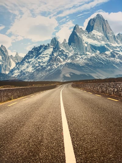 This media file depicts a winding asphalt road bordered by a stone wall on one side and majestic, snow-capped mountains on the other. The road features a white center line and intermittent yellow dashes along the edge. The mountains are rugged, with sheer rock faces and patches of snow, suggesting a high altitude. The sky is blue with scattered white clouds, indicating fair weather. The scene is oriented vertically, with the road and mountains appearing to ascend or descend. The lighting suggests it is daytime. There is no visible text in the image. The location is identified as Minzag, Russia.