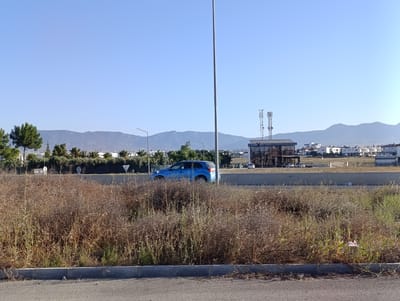 In North Nicosia, Northern Cyprus, a blue car is seen driving on a road. The surrounding area is dry and covered in brush. In the background, there are buildings and mountains. A tall pole stands in the foreground.