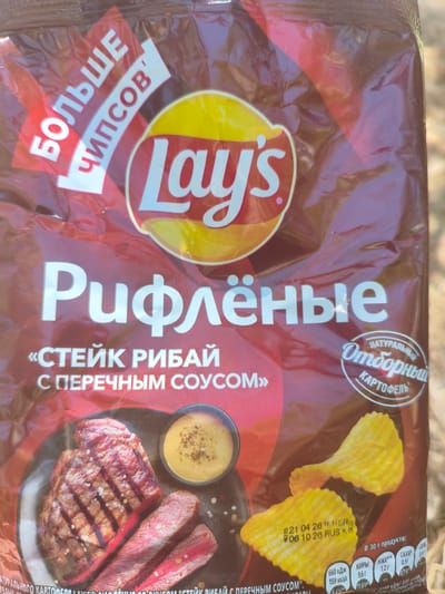 This is a close-up photograph of a bag of Lay's potato chips. The bag is primarily dark brown with a large red and yellow Lay's logo prominently displayed. The text on the bag is in Russian, and it indicates that the flavor is "Ribeye Steak with Pepper Sauce" («СТЕЙК РИБАЙ С ПЕРЕЧНЫМ СОУСОМ»). There is also text that reads "MORE CHIPS" (БОЛЬШЕ ЧИПСОВ) and "NATURAL SELECTED POTATOES" (НАТУРАЛЬНЫЙ ОТБОРНЫЙ КАРТОФЕЛЬ).

The image shows a depiction of a grilled steak with a side of sauce and two ruffled potato chips, suggesting the flavor profile. Nutritional information is also visible on the lower right side of the bag, along with a date code "21 04 26 18:15 SR 206 10 26 RUS K&" and "In 30g of product:" followed by values for calories, fats, carbohydrates, sugar, and salt. The lighting suggests it is daytime, possibly outdoors or near a window, as indicated by the warm ambient light and some environmental reflections on the plastic packaging. The overall impression is of a product advertisement. The location context provided is Alchevsk, Ukraine, suggesting this is where the product is being sold or photographed. There are no people visible in the image.