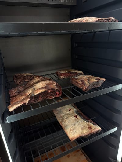 The image shows several cuts of beef dry-aging on metal racks inside a dark, enclosed unit with a bright light on the left side. The meat appears to be raw and has a dark, cured exterior. The unit has multiple shelves, and the meat is arranged on three of them. The top shelf has one piece of meat with a dark crust. The middle shelf has three pieces of meat, two of which are large cuts with visible marbling and a thick layer of fat. The bottom shelf has one large cut of meat covered in a thick layer of white fat. The interior of the unit is dark gray, and the racks are made of black metal. The exterior of the unit appears to be black plastic. The image was taken from a low angle, looking upwards into the unit. There is no indication of the location in the city of Dsegh, Armenia.