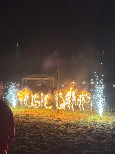 The image depicts an outdoor nighttime scene, likely at an event. In the foreground, a person wearing a maroon garment is partially visible on the left. The main subject is a large sign spelling out "MUSIC CAMP" that is engulfed in flames. The sign is constructed of metal letters and is supported by a metal frame. On either side of the flaming sign, there are pyrotechnic fountains, one on the left and another on the right, emitting sparks and light. In the background, there is a white tent with people gathered underneath it, and further back, there are more lights that might indicate buildings or other structures. The ground appears to be a mix of sand and grass. The sky is dark and appears to have some faint vertical streaks, possibly indicating rain or falling debris from the pyrotechnics. While the image does not contain specific visual cues to pinpoint the location to Menzelinsk, Russia, it captures a festive and dramatic event.
