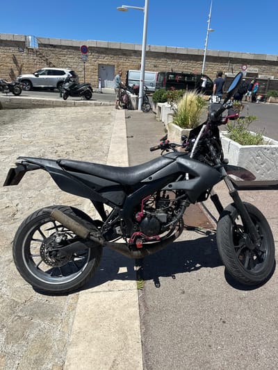A black supermoto motorcycle is parked on a cobblestone and asphalt surface in Saint-Tropez, France. The motorcycle is facing left in the frame. The front wheel is on the right side and the rear wheel is on the left. It has a black seat, a black body, and a black exhaust. Pink accents are visible on the engine and footpegs. The motorcycle is in the foreground.

In the background, there is a stone wall topped with a railing and planters. Several people are visible in the background, as well as a white SUV, a dark van, and a smaller motorcycle. The sky is clear and blue. The scene appears to be a street or pathway near a waterfront area.