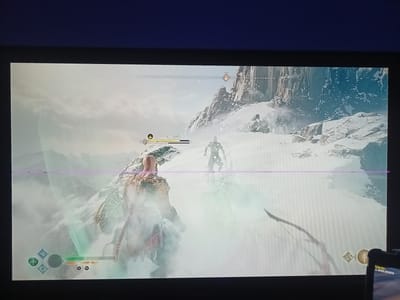 The image shows a video game scene in a snowy, mountainous environment. Two characters are visible. The foreground character is a muscular male figure with red hair, wearing a horned helmet and ornate armor. He is facing away from the viewer, toward another character in the middle ground. This second character appears to be a man with fair skin and short dark hair, wearing dark armor. They are standing on a snowy slope with a sheer cliff face in the background. The sky is overcast and hazy, with some mist or fog obscuring the distant landscape. A horizontal purple line runs across the middle of the screen, suggesting a display artifact. A game interface is visible at the bottom left, showing icons and a health bar. The bottom right of the screen shows what appears to be a controller with a small screen displaying the word "Цель" (which translates to "Target" or "Goal" in Russian) and "1.000.000$". There is no discernible connection to the city of Oberviechtach, Germany.