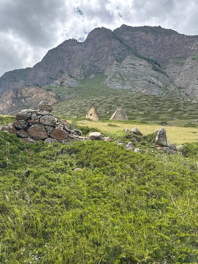 In El'tiubiu, Russia, a view captures ancient stone structures set against a dramatic mountain backdrop. In the foreground, a pile of weathered, reddish-brown stones, some with lichen growth, sits amidst lush green, low-lying vegetation and tall grasses. To the right, scattered rocks punctuate the grassy terrain. Further in the background, two distinct ancient stone structures are visible. One is a conical tomb with a small rectangular opening. Adjacent to it is a pyramid-shaped stone structure. The scene is framed by a massive, rugged mountain range with textured gray rock faces, streaks of green vegetation on its slopes, and a cloudy, overcast sky above.