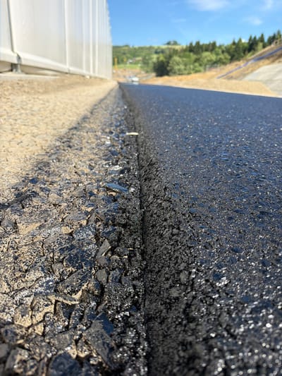 The image is a low-angle, close-up shot of a newly paved asphalt surface bordering a construction site. On the left, a rough, unpaved area with gravel and dirt is visible, adjacent to a white, tent-like structure. The dividing line between the unpaved area and the fresh asphalt is sharp. The asphalt surface is dark, wet, and reflects the sunlight, creating numerous small, bright highlights. In the background, a hill covered with trees and a clear blue sky can be seen. There are no people or vehicles prominently featured in the immediate foreground. The scene appears to be a section of road construction in an outdoor environment, possibly in a rural or suburban area, given the natural landscape in the background. The location is identified as Janowice, Poland.