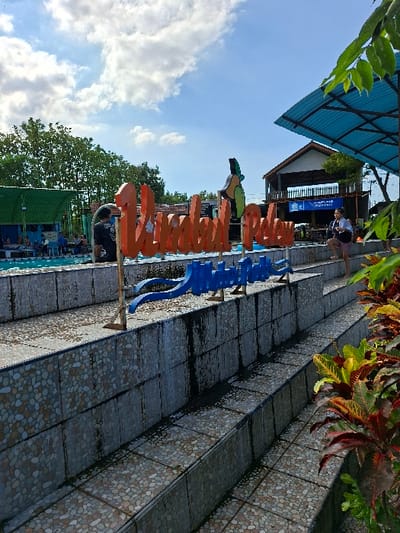 The image shows a tiled staircase leading up to a large orange sign that says "Umbul Pelow" in a pool area. The sign is next to a pool with blue water and several people are in the water. A person wearing a black shirt is standing by the pool, and another person wearing a white shirt is walking on the side of the pool. The background shows a lush, tropical green landscape with a blue sky with some white clouds. There is a blue awning above a building to the right of the sign. The scene is located in the Kecamatan Tulung, Indonesia at coordinates -7.592 latitude, 110.65 longitude.