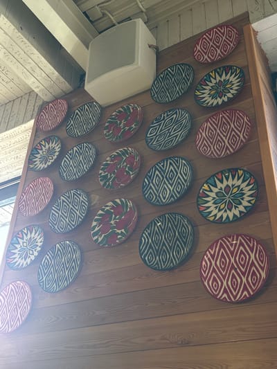 An interior shot shows a wooden wall adorned with numerous oval, decorative plates. The plates feature a variety of patterns, including geometric designs in red and white, blue and white, and colorful floral or starburst motifs. A white speaker is mounted on the wall above the plates. The ceiling, made of wooden beams and planks, is visible at the top of the image. The scene appears to be inside a building, possibly a restaurant or home, in the city of Solnechnoye, Russia, judging by the style of the interior elements and the context provided.