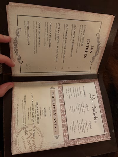 The image displays an open menu, held by a person's hands, in what appears to be a dimly lit restaurant setting. The background is dark and out of focus. The menu has a dark brown cover and two light-colored pages with decorative borders.
The left page is titled "LES ENTRÉES" (Starters) and lists several French dishes with their prices in euros. Items include "SOUPE À L'OIGNON" (French onion soup), "ESCARGOTS EN PERSILLADE #9" (Burgundy snails), "OS À MOELLE GRATINÉ AU PARMESAN" (Roasted marrowbones), "PÂTÉ EN CROÛTE, SALADE, PICKLES", "GRAVLAX DE SAUMON", "OEUFS EN MEURETTE" (Poached egg in wine sauce), and "HUÎTRES SPÉCIALES" (Special Oysters). Prices range from 19 to 38 euros.
The right page is divided into two main sections: "Les Salades" and "POUR LES ENFANTS" (For Children). The salads offered are "SAVOYARDE" (with Reblochon and goat cheese), "VIKING" (with tuna tataki, salmon gravlax), and "VEGGIE" (with falafel and roasted vegetables), priced at 33, 34, and 31 euros respectively. The children's menu, for those up to 10 years old and priced at 28 euros, includes a beverage, a choice of "STEAK HACHÉ FRITES" (Minced beef with French fries) or "CASSOLETTE DE RAVIOLES AUX CÈPES" (Cassolette of porcini ravioli), and a dessert of "bouette (mousse au chocolat)" or vanilla ice cream.
A watermark on the right page reads "La Peau de Vache Val d'Isère," indicating the restaurant's name and its location in Val-d'Isère, France. The bottom of the menu states "prix nets, en euros * service compris" (net prices, in euros * service included).