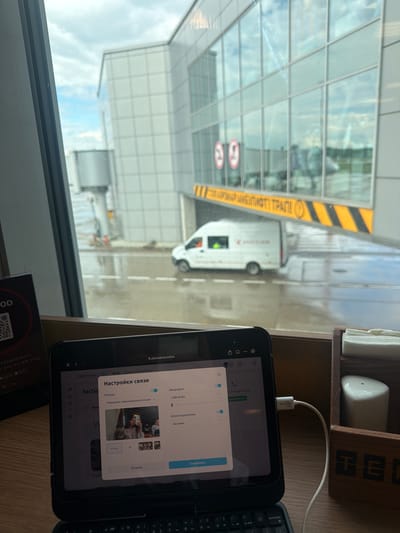 An interior view from an airport waiting area shows a tablet computer displaying a video conferencing interface, with the tablet placed on a keyboard. Outside the window, an airport jet bridge and a white service van are visible, with the ground appearing wet. In the foreground, a wooden napkin holder and a salt shaker are present. The tablet displays "Настройки связи" (Connection Settings) with options for Camera, Microphone, and Sound. The webcam preview shows a woman taking a selfie. The overall setting suggests a traveler using technology while waiting for a flight.