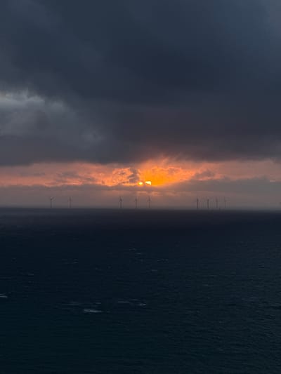 The image captures a dramatic sunset or sunrise over an offshore wind farm near Fécamp, France. The scene is divided horizontally, with the dark, textured sea occupying the bottom half and a vibrant, cloudy sky above.
A long line of offshore wind turbines extends across the horizon, silhouetted against the bright sky. The sun, a brilliant orange and yellow, is partially visible, peeking through a break in the clouds, casting a warm, intense glow. The clouds directly surrounding the sun are illuminated with striking oranges and yellows, transitioning to lighter greys and then darker, heavier greys and blues towards the left side of the sky. This contrast creates a dynamic and impactful sky.
The sea below is a deep, dark blue or almost black, with subtle textures indicating gentle waves or surface ripples, reflecting the low light. There are no people or other objects besides the wind turbines. The time of day is clearly dawn or dusk, characterized by the sun's low position and the vivid sky colors. The atmosphere is powerful and serene, showcasing both natural beauty and human engineering. No visible text is present in the image.