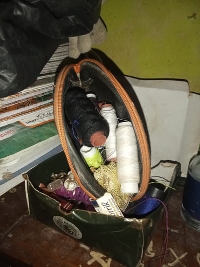The image displays an indoor scene on a wooden surface within a location in Ciputri, Indonesia. In the center, an open, upright, tan-orange zippered fabric pouch contains several spools of thread (black, white, and lime green), a gold sequined trim, and other small, unidentified sewing accessories. Directly below this pouch, a dark green, partially crushed cardboard box holds various items including a clear plastic bottle cap, purple coiled material, and a white and blue package labeled "FOTTIK DOUBLE EDGE RAZOR."
To the immediate left of the central items, a stack of books or notebooks is visible. The top visible book is green with white text, partially legible as "Sekolah: MTs Al-J" and "KARYA DUYA." A crumpled black plastic bag rests on the uppermost book, beneath which a light grey, possibly fabric or stuffed, object is partially visible. The stack of books is positioned against a vertical, light olive-green wall.
To the right of the central objects, a clear glass bottle with a dark blue label is partially visible. Behind it, a light-colored, possibly white, folded piece of cardboard stands against the wall. The underlying surface is a dark brown wood, exhibiting faded, light-colored patterns or scratches. No individuals are present in the frame.