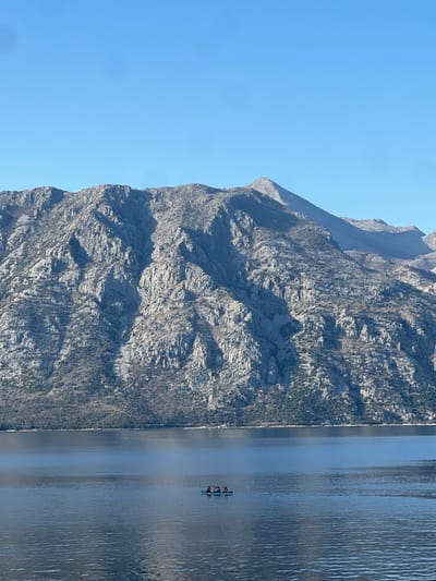 A panoramic view captures a vast body of water at the base of imposing, rugged mountains under a clear, azure sky. The location is Bogdašići, Montenegro. The mountains, occupying the upper two-thirds of the frame, are characterized by grey, rocky, and deeply eroded surfaces with sparse, scrubby vegetation in shades of brown and dark green. Deep shadows indicate strong sunlight, highlighting the craggy textures and valleys. A prominent, pointed peak rises on the upper right side of the mountain range. The water, a tranquil expanse of deep blue, occupies the lower third of the image, showing minimal ripples and reflecting the sky's brightness. Centered horizontally in the mid-ground of the water, a small, dark-colored vessel, possibly a kayak or inflatable boat, contains three human figures. Two figures are seated near the front, with a third behind them. One individual exhibits an orange or red element on their upper torso, consistent with a life vest or clothing. No distinct actions are discernible from the individuals or the vessel due to the distance. The shoreline at the base of the mountains is dimly visible, appearing as a thin strip of darker land with sparse trees or bushes.