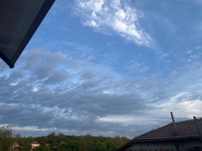The image depicts a partially cloudy sky above the city of Morava, Bulgaria. The sky is predominantly blue with scattered white and grey clouds. A section of a building overhangs the top left corner of the frame, obscuring the sky. Below the horizon line are trees and rooftops, including a building with a brown tile roof and two exposed pipes. The trees appear to be part of a forest or wooded area. Several small buildings are visible in the distance. A small, dark object, possibly a bird, is visible in the sky.