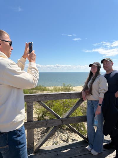In the city of Morskoye, Russia, three people are on a wooden boardwalk overlooking a beach and the sea. A man in the foreground on the left is wearing sunglasses and a cream-colored hoodie and is holding up a smartphone, appearing to take a picture. To the right, a woman wearing a beige jacket, blue jeans, and a dark baseball cap stands next to a man in a dark t-shirt and a camouflage baseball cap. The woman is holding a bottle of water in her right hand. The sea is calm and a light blue, with a sandy beach in the distance. The sky is clear and blue with a few scattered white clouds. The boardwalk is made of weathered wood and has a railing with a geometric design. Green foliage is visible on the sand dunes.