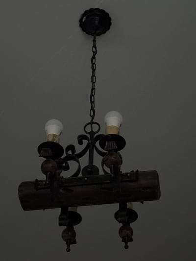 This media file shows a rustic-style chandelier hanging from a ceiling. The chandelier features a central wooden beam from which multiple light fixtures extend. Each light fixture consists of a metal holder with a bulb. Two bulbs are clearly visible, appearing to be energy-efficient or LED bulbs. The metalwork of the chandelier is dark and ornate, with curved decorative elements. A chain connects the chandelier to a decorative ceiling mount on the left side of the image. The overall style suggests a rustic or traditional decor. The background is a plain, gray ceiling. There is no activity or event taking place; the image is a still shot of the object. No people, text, or specific time-of-day or weather cues are visible. The setting appears to be indoors, likely a room within a home or building. The location context provided is Sant'Angelo di Piove di Sacco, Italy, but this cannot be confirmed from the visual information alone.