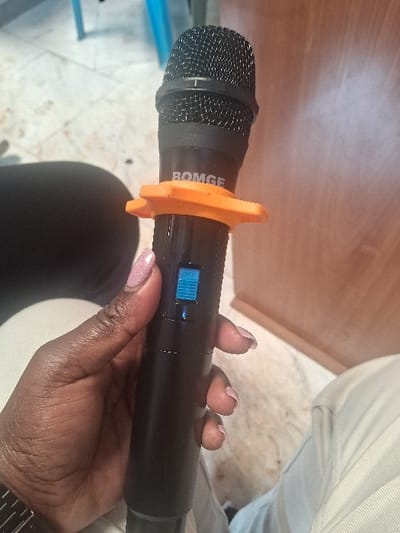 A person's right hand, with dark skin and light pink manicured nails, holds a black wireless BOMGE microphone vertically. An orange, star-shaped anti-roll ring is present below the microphone head. A blue "OFF" switch with a blue indicator light is visible on the microphone's body. A silver-colored watch with a metal band is on the person's wrist. The individual is wearing light-colored trousers, visible on the right. The background features a light-colored tiled floor, a wooden furniture piece on the right, and a dark fabric object on the left. The scene is indoors, located in Muthambi ward, Kenya.