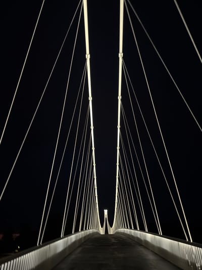 The media file presents an abstract, architectural view of a modern bridge or structure at night, set against a stark, black background. The central elements are two parallel, brightly illuminated horizontal beams or pipes. From these beams, numerous thin, light-colored cables or suspension lines extend diagonally upwards and downwards, creating a symmetrical, fanned pattern across the dark expanse. Each beam features regularly spaced connectors from which these cables originate.
On the far right side of the image, a large, curved section of the structure's wall or side is visible, brightly lit along its contours and textured surface. This surface appears to have a ribbed or slatted design, which creates distinct patterns of light and shadow, emphasizing the curve and material. The overall scene is devoid of any specific activity, focusing solely on the static, illuminated architecture.
The time of day is clearly night, as indicated by the deep black background and the reliance on artificial lighting to define the structural elements. The crispness of the light and lines suggests clear weather. While the image itself doesn't contain explicit location cues, the provided context identifies the location as Osijek, Croatia, suggesting this is a modern public infrastructure, likely a pedestrian or cable-stayed bridge. No text is visible in the image.
