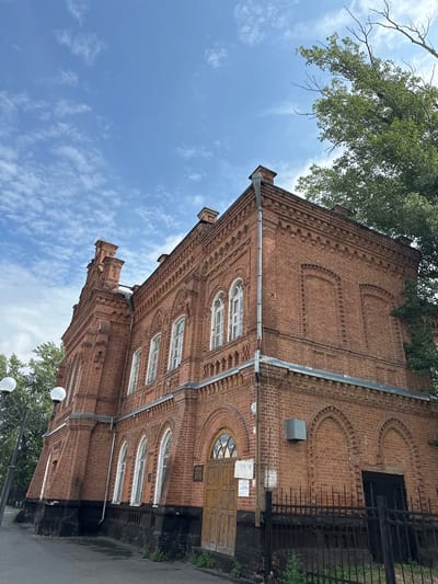 An exterior low-angle shot shows a large, ornate brick building against a bright blue sky with scattered clouds. The building is constructed of red brick with decorative elements, including arched windows and intricate brickwork along the roofline and around the window frames. A tall, pointed architectural feature adorns the left side of the building. To the right, a large tree with green foliage partially obscures the building. In the foreground, a paved walkway runs alongside the building, with a black metal fence bordering it. A lamppost with two globe-shaped lights is visible on the left. The building appears to be in a state of disrepair in some areas, with visible water damage at the base and faded paint. A wooden door is set into an arched entryway on the right side of the building, with a sign attached to the wall next to it.