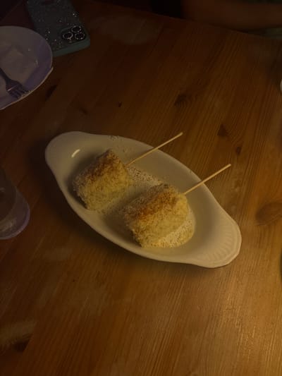 Two pieces of corn on the cob, each with a skewer inserted, are presented on a white oval dish. The corn appears to be coated in a light-colored crumb or cheese mixture and sprinkled with red seasoning. The dish is placed on a wooden table. To the left of the dish is a partially visible glass of water and a white plate with a fork. In the upper left corner of the image is a glittery teal phone case. The scene is dimly lit, suggesting an indoor setting, possibly a restaurant. The image does not provide any specific information to determine the location within San Juan, United States of America.