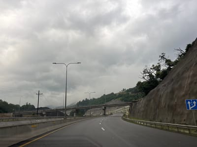 The image shows a view from a car driving on a highway in Tsakva, Georgia, on a cloudy day. The highway curves to the right, with a steep, rocky embankment on the right side. There are guardrails along the edge of the road and a traffic sign indicating that the lane splits into three directions, with two going straight and one turning left. In the distance, a bridge spans over the highway, and a power line with a cross-shaped pole is visible. Lush green trees and hills are present on either side of the highway. The sky is filled with heavy, grey clouds, suggesting overcast weather.