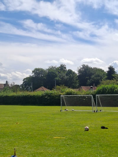 The image depicts a green, manicured sports field, likely a football pitch, in Richmond, United Kingdom. Two football goals with white netting are visible on the right side of the frame. Several footballs are scattered on the grass, with one prominently placed in the foreground. A black object, possibly a pair of gloves, lies near the goal. Yellow markers are also visible on the field.
The background features a line of mature trees and dense green hedges, with glimpses of red-tiled rooftops of houses peeking through the foliage. The sky is a bright blue with wispy white clouds, suggesting a clear and sunny day, likely during daylight hours. The overall atmosphere is serene and peaceful, with no people visible in the frame. The setting appears to be a well-maintained outdoor facility, typical of a park or sports club. There is no visible text in the image.