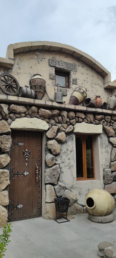 This is a medium-shot, eye-level, exterior, vertical, full-color photograph of a building facade that appears to be an old or stylized winery or rustic establishment in Voskevaz, Armenia. The building is constructed with a rough, textured, light-colored stucco and incorporates rough-hewn stones of various sizes for accentuation and structural elements.
The main focus is on the entrance, a heavy wooden door with dark metal decorative hinges and a central ornate handle. The door is framed by large, irregular stones. To the left of the door, a wooden cartwheel and a small wooden barrel are displayed on a stone ledge. Above this ledge, there are several antique-looking pottery vessels of varying shapes and sizes, including a tall, narrow vase with rope detailing, and a large, round, tan-colored clay pot with a wide opening, resting on a stone base. A metal watering can or jug is also present on the ledge.
To the right of the door, a small, wooden-framed window with a diamond-patterned pane is visible. This window is also framed by the textured stucco and stones. A black metal trash receptacle with decorative detailing and a plastic liner is placed on the paved ground in front of the building, near the door and the large clay pot. The paving stones are a light gray color. The sky is overcast and pale blue. A small, leafy green plant is visible in the lower left corner of the frame.