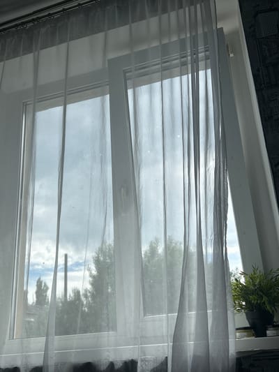 The image captures an indoor perspective of a partially open window in Chernomurovskii, Russia. Sheer white curtains hang across the window, diffusing the bright daylight entering the room and gently billowing, suggesting a light breeze.

The window itself is a modern type, with the top section tilted inward, facilitating ventilation. Through the translucent curtains and the window panes, a mostly cloudy sky is visible, with patches of brighter white indicating daylight. Green foliage from trees can be seen outside, partially obscured by the curtains and the cloudy atmosphere, indicating an outdoor natural setting.

Above the window frame, a small potted plant with green leaves sits on an internal ledge. To the right of the window, partially visible through the curtains and reflected light, are some jars or containers, possibly on a shelf. The indoor environment also shows a dark, textured surface along the top edge of the frame, likely part of the ceiling or an adjacent wall. The time of day appears to be daytime, likely mid-day or afternoon, under a cloudy sky. No discernible text is visible on any objects in the image.