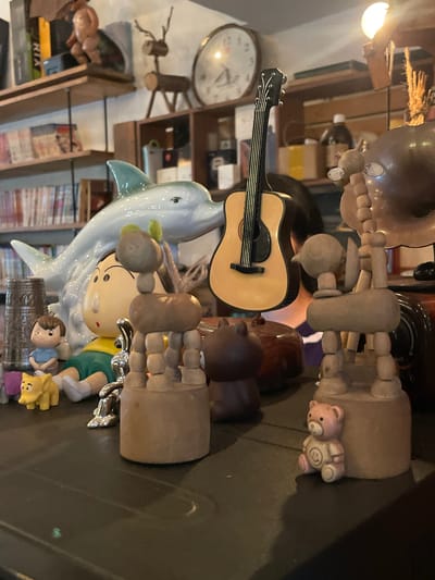 The image depicts a cluttered display of various decorative items on a dark, reflective surface, likely a countertop or shelf. In the foreground, a small, pink teddy bear with a smiley face on its shirt sits to the right. Behind it, to the left, a yellow figurine of a piglet is positioned near a doll wearing a blue shirt and green shorts. A silver-colored, abstract sculpture is situated between these figures. In the mid-ground, a light blue ceramic dolphin is prominently featured, with its head pointing to the left. A miniature acoustic guitar with a light-colored body and black strings stands upright behind the dolphin. To the right of the guitar, several wooden sculptures are visible, including a figure resembling a dog or deer with articulated limbs and a round head. Further to the right, a dark, metallic object, possibly part of a gramophone or similar device, is partially visible. The background reveals shelves filled with books and various other objects, including a large clock on the wall and a decorative wooden reindeer figure. The lighting suggests an indoor environment, possibly a shop or a decorated living space. The location is identified as Liên Nghĩa, Vietnam.