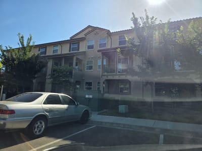The image depicts a multi-story building, likely an apartment complex or townhouses, under a bright, sunny sky. The building's exterior is a neutral beige color with a red tile roof. Various windows, some with dark shutters, are visible on its facade, along with covered balconies and supporting columns. Trees with green foliage are present in the foreground and surrounding the building, casting shadows and partially obscuring the structure due to the harsh sunlight.
In the foreground, a silver sedan is parked on the left side of the frame. The car appears to be older, with some visible wear and tear. The scene is set on a paved area, possibly a parking lot or street, with a concrete curb and sidewalk. Behind the car and in front of the building, there is a large green utility box. Another smaller, lighter green utility unit is visible on the lawn area to the right of the sidewalk.
The time of day appears to be late morning or early afternoon, given the high angle of the sun and the strong sunbeams and lens flare cutting across the image, suggesting a clear and sunny day. The location context of Elk Grove, USA, places this scene in a suburban or residential environment. There is no discernible activity or event taking place; it is a static scene. No emotions are conveyed.
Visible text includes labels on the green utility box, which are not clearly legible but appear to be safety or identification markings. There is also text on the side of the silver car, likely a model badge, but it is too blurry to read.