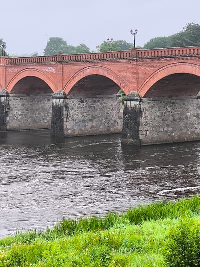 The image shows a red brick arched bridge spanning across a river in Kuldīga, Latvia. The bridge has stone pillars and is topped with a railing and two streetlights. The river is dark and slightly choppy, with green grass lining the bank in the foreground. Trees are visible in the background, and the sky is overcast.