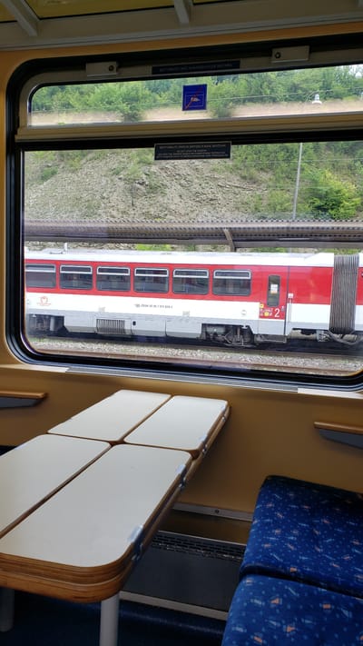 The image shows the interior of a train carriage at the Kraľovany, Slovakia train station. Through the window, a red and white passenger train is visible on the tracks, parked at the station platform. The window area features warning signs, one in Slovak and German, indicating that opening the window will disable the air conditioning. Inside the carriage, two folding tables are set up, positioned over a dark blue seat with a patterned fabric. The tables are a light color with rounded edges and are supported by white legs. The overall lighting suggests it is daytime.