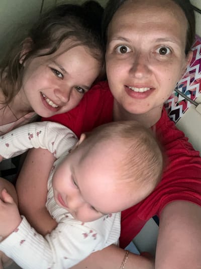 A mother and her two daughters are posing for a selfie. The mother is wearing a red shirt and is holding a baby in her arms. Her older daughter is standing next to her, smiling at the camera. The baby is looking down at the mother's chest. The background is blurred, but it appears to be a domestic setting. The image was taken in the city of Mozhga, Russia.