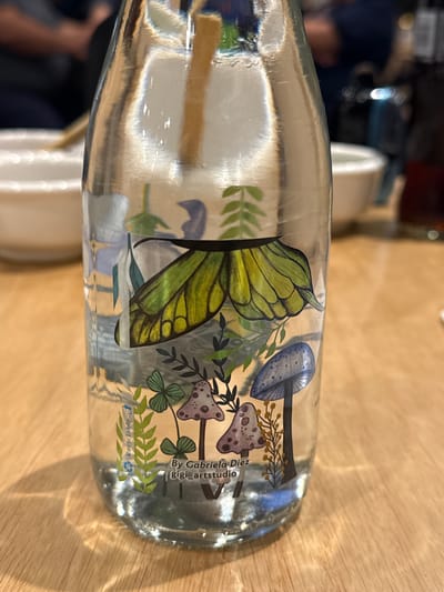 The image depicts a clear glass bottle decorated with a botanical illustration of mushrooms and a butterfly. The illustration also includes the text "By Gabriela Diez Gigi_artstudio" and a recycle symbol. The bottle is resting on a light-colored wooden surface. The photograph was taken indoors. The location, Querétaro, Mexico, is not visually confirmed in the image.